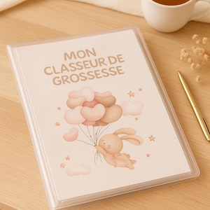 Peut inclure: Un classeur en plastique transparent avec le texte "MON CLASSEUR DE GROSSESSE" sur la couverture. La couverture présente un lapin de dessin animé tenant des ballons. Une tasse de thé et un stylo doré sont sur la surface en bois.