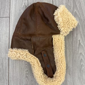 May include: A brown leather aviator hat with cream-coloured shearling lining and ear flaps. The hat has a button closure on the side. Designed to provide warmth and protection from the elements.