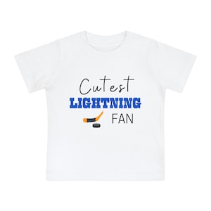 May include: White toddler t-shirt with the text "Cutest Lightning Fan" in blue and black. A hockey stick and puck are also included in the design.