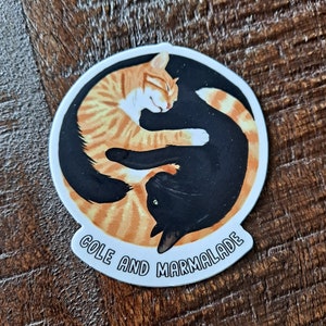 May include: A round sticker with a yin-yang design featuring two cats curled up together. The orange cat is on the left and the black cat is on the right. The text "Cole and Marmalade" is written in a white arc around the bottom of the sticker.