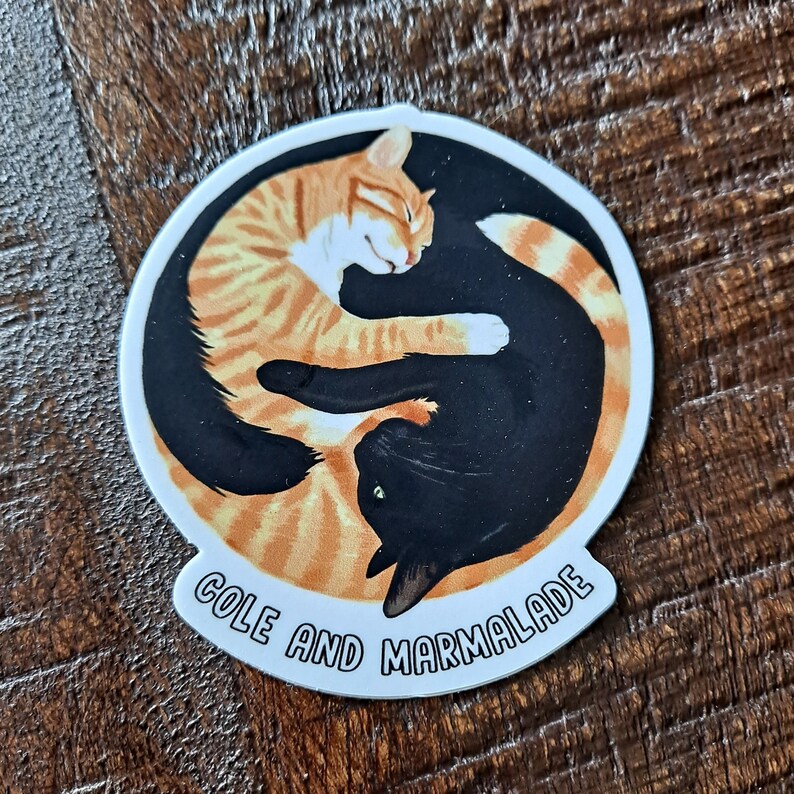 May include: A round sticker with a yin-yang design featuring two cats curled up together. The orange cat is on the left and the black cat is on the right. The text "Cole and Marmalade" is written in a white arc around the bottom of the sticker.