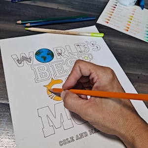 May include: A coloring page with the text "World's Best Mom" with a star and a globe. A hand is coloring the star with an orange pencil.