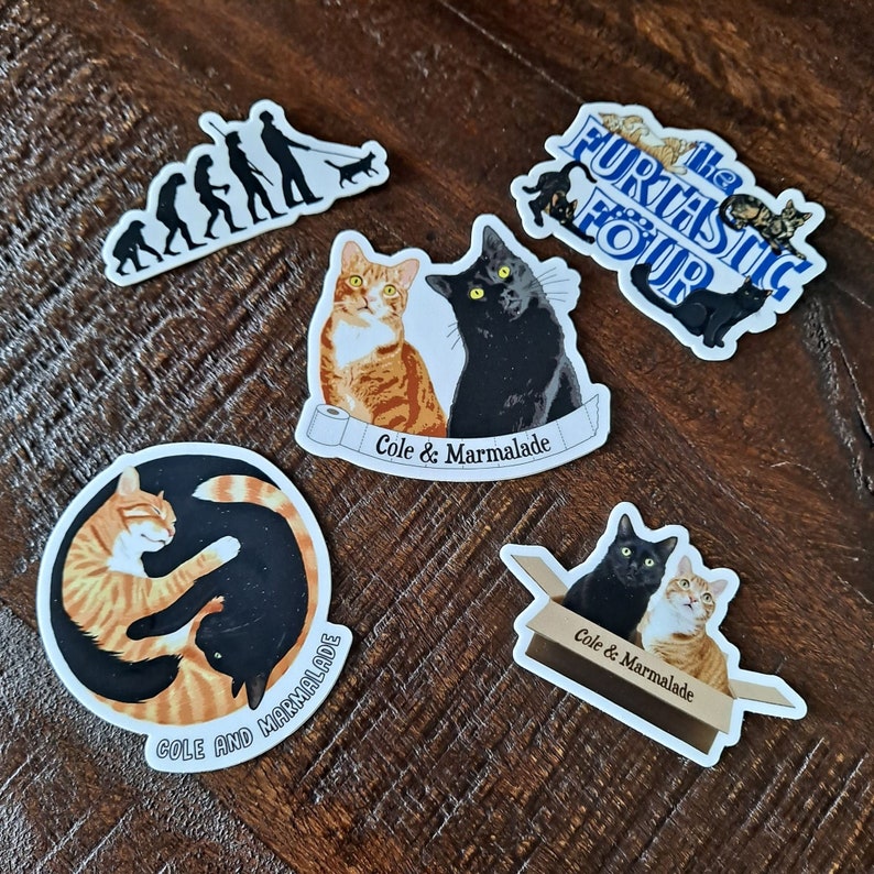 May include: Four stickers featuring cats. One sticker shows two cats, one orange and one black, with the text "Cole & Marmalade". Another sticker shows two cats, one orange and one black, in a box with the text "Cole & Marmalade". A third sticker shows two cats, one orange and one black, curled up together with the text "Cole and Marmalade". The fourth sticker shows a group of cats with the text "The Fur-tastic Four".