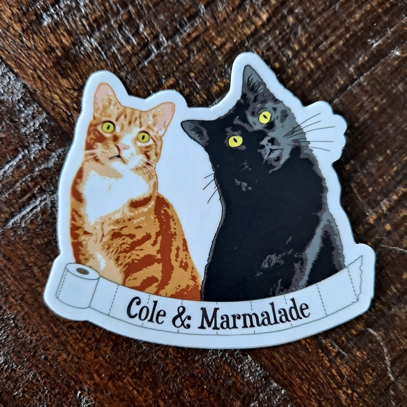 May include: A white sticker with a black cat and an orange tabby cat. The cats are looking at the camera. The sticker has a white banner with the text "Cole & Marmalade" on it.