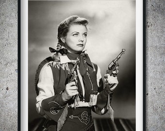 May include: A black and white photo of a woman wearing a western-style outfit and holding two pistols. She is wearing a white shirt with a brown vest and a bandana around her neck. She is looking to the right of the frame.