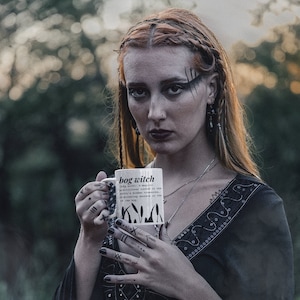 Puede incluir: Una mujer con el pelo largo y rojo oscuro sostiene una taza de cerámica blanca con texto e ilustraciones negras. La taza dice "bog witch (bog wɪtʃ): A magical practitioner rooted in the earth's hidden treasures, discovering secrets in the bog."