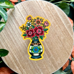May include: A colorful sticker featuring a vase with a floral arrangement. The vase is blue and white with a yellow background. The flowers are red, pink, and white.