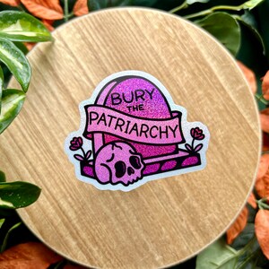 May include: A glittery pink and purple sticker featuring a tombstone with the text "BURY THE PATRIARCHY" written on a banner across the front. A skull sits at the base of the tombstone, flanked by two small flowers on either side of the design.