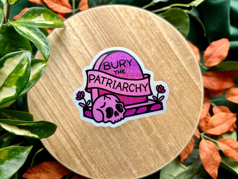 May include: A glittery pink and purple sticker featuring a tombstone with the text "BURY THE PATRIARCHY" written on a banner across the front. A skull sits at the base of the tombstone, flanked by two small flowers on either side of the design.