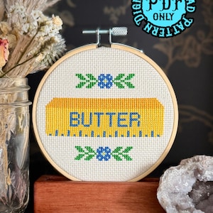 May include: A cross-stitch embroidery hoop featuring a yellow butter stick design with the word "BUTTER" in blue. The design is framed by green and blue floral accents. The text "DIGITAL DOWNLOAD PDF ONLY" is in a blue circle.