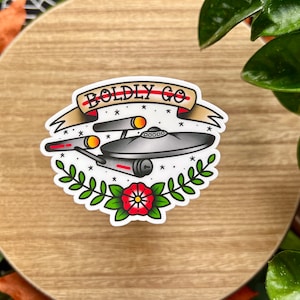Peut inclure: Un autocollant blanc avec une illustration noire et rouge d'un vaisseau spatial avec le texte "BOLDLY GO" dans une bannière au-dessus. Le vaisseau spatial est entouré d'étoiles et d'une fleur rouge.