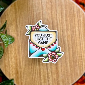 May include: A sticker featuring a tattoo-style design of an envelope with the text "YOU JUST LOST THE GAME." The envelope is teal with a red heart and floral accents. The sticker is placed on a wooden surface.