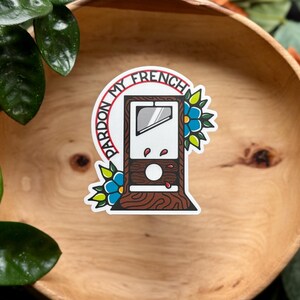 May include: A die-cut sticker featuring a brown wood-grain guillotine with a silver blade and red blood droplets. The text PARDON MY FRENCH is arched over the top in red letters, flanked by two blue flowers with green leaves on either side.