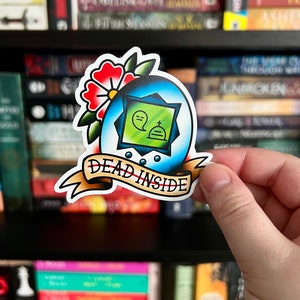 May include: A die-cut sticker featuring a blue egg-shaped frame with a green center containing a ghost and tombstone icon. Above the egg is a red flower with green leaves. A tan banner at the bottom displays the text DEAD INSIDE in black ink.