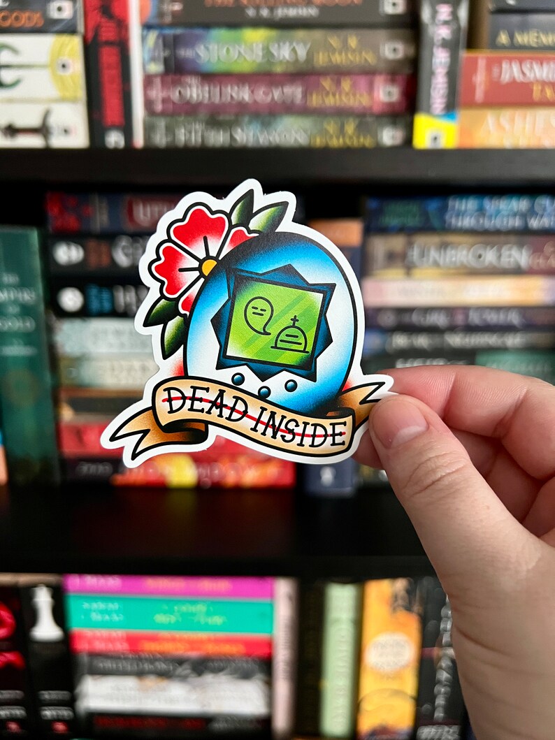 May include: A die-cut sticker featuring a blue egg-shaped frame with a green center containing a ghost and tombstone icon. Above the egg is a red flower with green leaves. A tan banner at the bottom displays the text DEAD INSIDE in black ink.