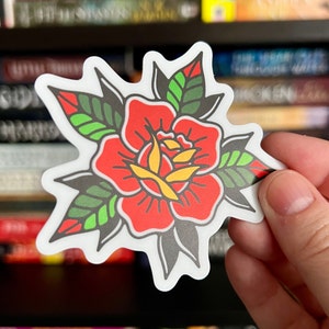 Pu&ograve; includere: Un adesivo fustellato raffigurante una rosa stilizzata con petali rosso vivo, un centro giallo e foglie verde scuro con riflessi verde chiaro. Il disegno &egrave; delineato da spessi tratti neri ed &egrave; rifinito con un bordo bianco netto e definito.