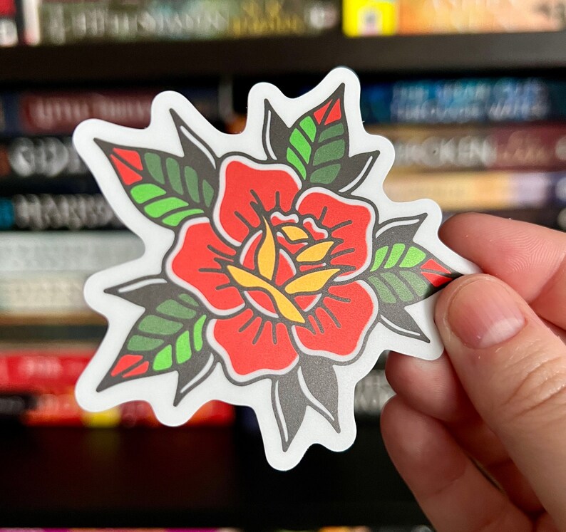 Pu&ograve; includere: Un adesivo fustellato raffigurante una rosa stilizzata con petali rosso vivo, un centro giallo e foglie verde scuro con riflessi verde chiaro. Il disegno &egrave; delineato da spessi tratti neri ed &egrave; rifinito con un bordo bianco netto e definito.