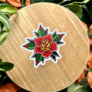 Pu&ograve; includere: Un adesivo fustellato con un'illustrazione di una rosa in stile tatuaggio tradizionale. Il fiore presenta petali rosso vivo con centri gialli, circondati da foglie verde scuro e nere, il tutto delineato da un bordo bianco marcato.