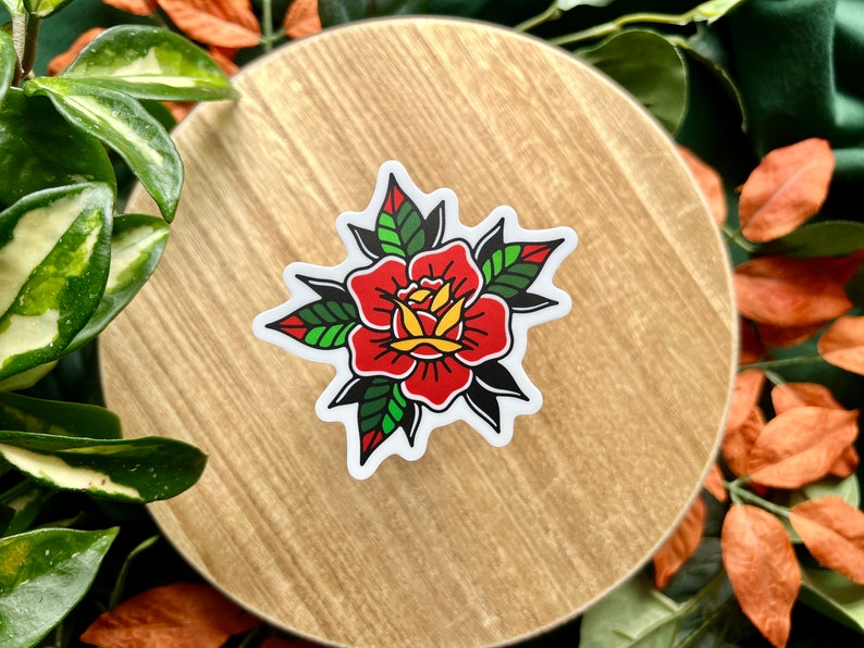 Pu&ograve; includere: Un adesivo fustellato con un'illustrazione di una rosa in stile tatuaggio tradizionale. Il fiore presenta petali rosso vivo con centri gialli, circondati da foglie verde scuro e nere, il tutto delineato da un bordo bianco marcato.