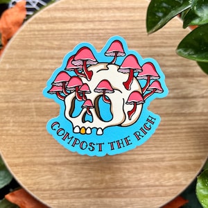 May include: A blue sticker with a white skull with pink mushrooms growing out of the top. The text "COMPOST THE RICH" is written in a curved arc below the skull.