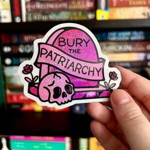 May include: A holographic sticker featuring a pink tombstone with the text "BURY THE PATRIARCHY" on a banner. A pink skull sits at the base of the tombstone, flanked by two small pink flowers on either side. The design has a glittery finish.