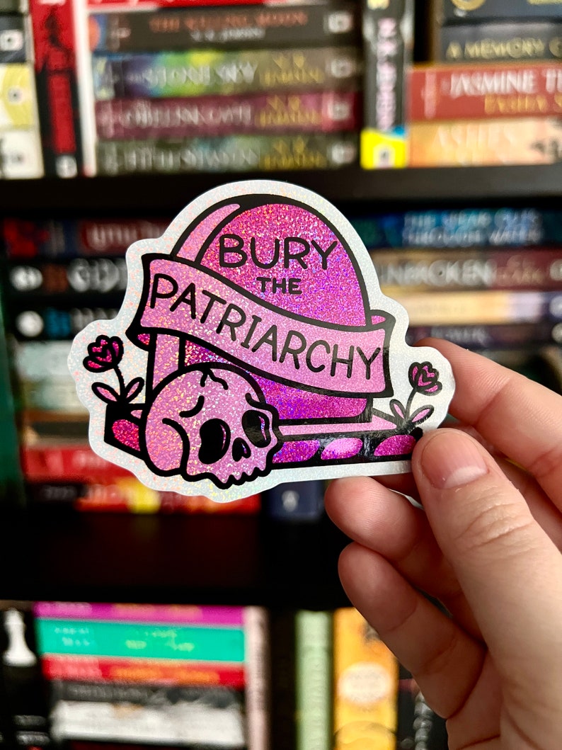 May include: A holographic sticker featuring a pink tombstone with the text "BURY THE PATRIARCHY" on a banner. A pink skull sits at the base of the tombstone, flanked by two small pink flowers on either side. The design has a glittery finish.