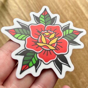 Pu&ograve; includere: Un adesivo fustellato che presenta un'illustrazione di una rosa stilizzata con spessi contorni neri. Il fiore ha petali rosso vivace, un centro giallo e foglie verdi con accenti rossi, il tutto su uno sfondo con bordo bianco.