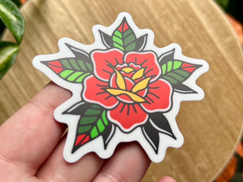 Pu&ograve; includere: Un adesivo fustellato che presenta un'illustrazione di una rosa stilizzata con spessi contorni neri. Il fiore ha petali rosso vivace, un centro giallo e foglie verdi con accenti rossi, il tutto su uno sfondo con bordo bianco.