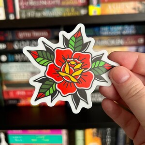 Pu&ograve; includere: Un adesivo fustellato raffigurante una rosa in stile tatuaggio tradizionale con petali rosso vivo, un centro giallo e foglie verde scuro con contorni neri. Il design &egrave; simmetrico con un bordo bianco e linee grafiche nette e ben definite.