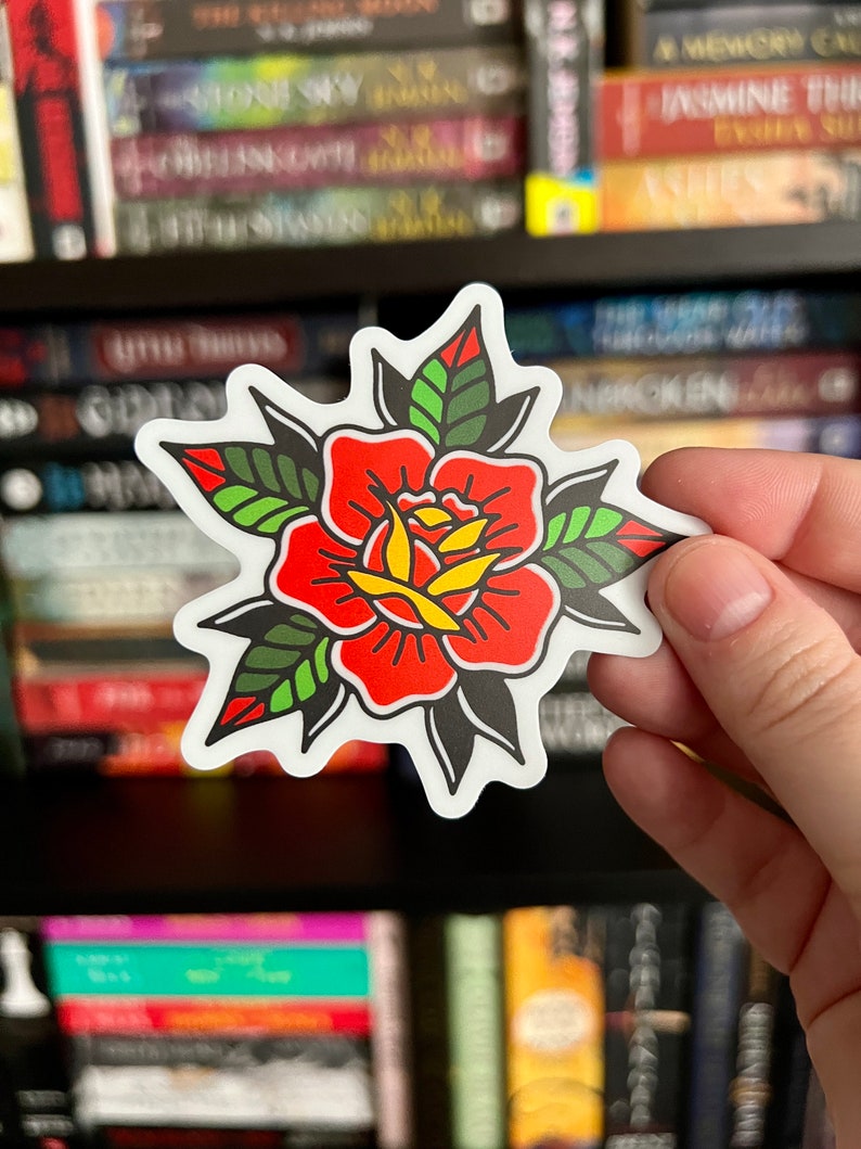 Pu&ograve; includere: Un adesivo fustellato raffigurante una rosa in stile tatuaggio tradizionale con petali rosso vivo, un centro giallo e foglie verde scuro con contorni neri. Il design &egrave; simmetrico con un bordo bianco e linee grafiche nette e ben definite.
