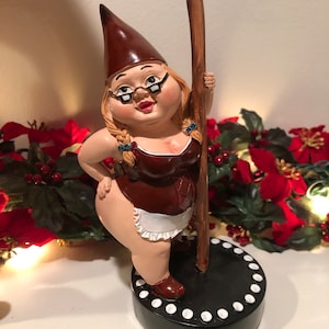 May include: A brown and white gnome figurine wearing glasses and a brown dress with a white apron. The gnome is holding a brown stick and standing on a black base with white dots.