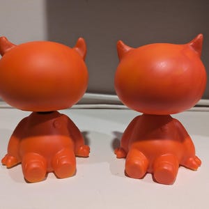May include: Two orange devil-shaped bobbleheads with horns and a rounded head. The bobbleheads are facing opposite directions.