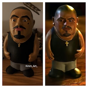 May include: A 3D printed figurine of a person with a black tank top, gray pants, and a gold chain with a cross pendant. The figurine has a brown complexion, a black goatee, and a black mustache. The figurine is standing on a wooden surface.