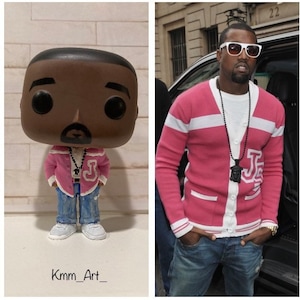 Puede incluir: Una figura Funko Pop personalizada de una persona que lleva un cárdigan a rayas rosa y blanco con un collar de colgante negro y blanco. La figura lleva pantalones vaqueros azules y zapatillas blancas. La figura está de pie sobre una superficie blanca. El fondo es una pared de ladrillo.