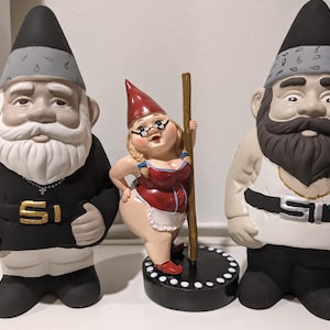 May include: Three garden gnomes, one male with a black outfit and a white beard, one female with a red dress and a pole, and one male with a white tank top and a black belt. All gnomes have gray hats with a black and white pattern.