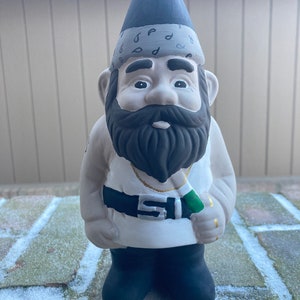 May include: A white garden gnome wearing a black bandana with a paisley pattern, a black shirt with a gold chain, a black belt with a silver buckle, and black pants. The gnome is holding a green object in its right hand.