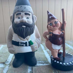 May include: Two garden gnomes, one wearing a bandana and holding a bottle, the other wearing a brown dress and holding a pole.