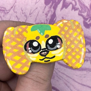 May include: A hand-painted yellow and orange waffle-textured pin featuring a stylized animal face with large black eyes, a small nose, and a green leaf-like detail on the forehead. The object has a flat, irregular shape with a grid pattern.