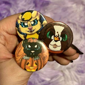 Puede incluir: Tres botones redondos y coloridos con diseños de animales de dibujos animados. Un botón presenta una criatura felina amarilla y azul con ojos azules. Otro muestra un animal marrón y blanco con ojos verdes. El tercer botón tiene un gato negro en una calabaza naranja.