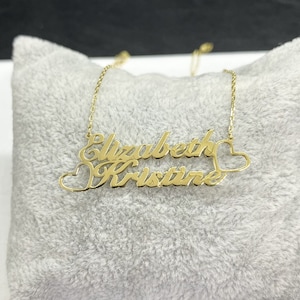 May include: A gold-colored necklace featuring the names "Elizabeth" and "Kristine" in a cursive font. The necklace includes heart-shaped accents on each side of the names and hangs on a delicate chain. The jewelry is displayed on a gray, plush surface.