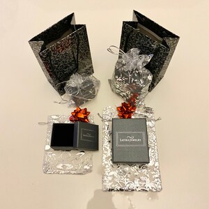May include: Two black gift bags with silver floral patterns, two small black gift boxes with red bows, and two silver gift bags with red bows and black boxes with the text "Latika Jewelry".