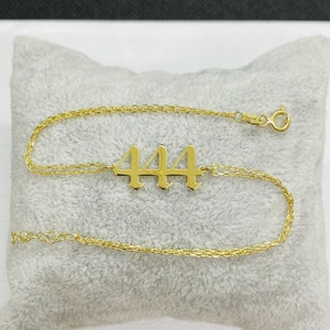 May include: Gold chain bracelet with a 444 charm. The chain is a delicate, double-strand design. The charm is in a bold, gothic font.