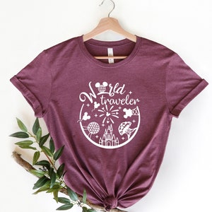 May include: A maroon t-shirt with rolled-up sleeves, featuring a white graphic that says "World Traveler" with iconic travel-themed illustrations. The design includes a Mickey Mouse head, fireworks, and a castle.