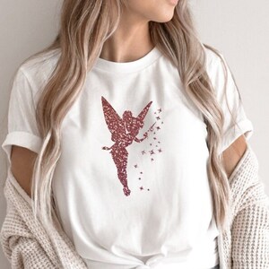 Puede incluir: Una camiseta blanca con un dise&ntilde;o de hada en purpurina rosa y estrellas. El hada sostiene una varita m&aacute;gica y tiene alas. La camiseta est&aacute; anudada a la cintura y la modelo lleva pantalones vaqueros azules y una chaqueta de punto crema.