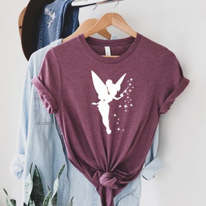 Puede incluir: Una camiseta color granate con una silueta blanca de Campanilla, el hada de Disney, esparciendo polvo de hadas.