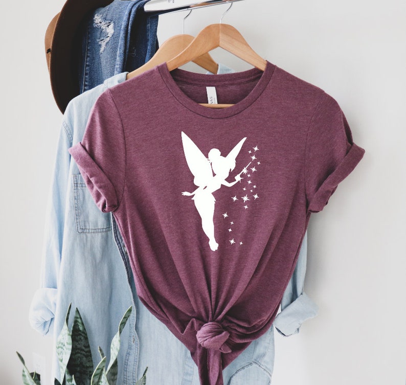Puede incluir: Una camiseta color granate con una silueta blanca de Campanilla, el hada de Disney, esparciendo polvo de hadas.