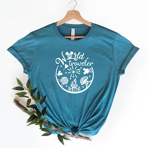 May include: Teal t-shirt with a white graphic featuring the text "World Traveler" and iconic imagery. The design includes a Mickey Mouse head, fireworks, a castle, and other travel-themed elements. The shirt is displayed on a wooden hanger.