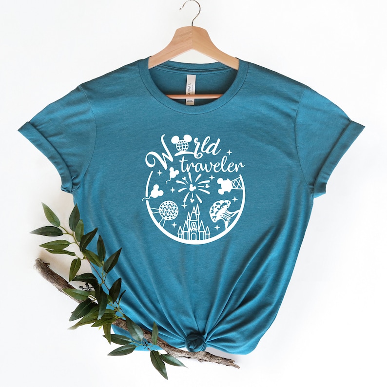 May include: Teal t-shirt with a white graphic featuring the text "World Traveler" and iconic imagery. The design includes a Mickey Mouse head, fireworks, a castle, and other travel-themed elements. The shirt is displayed on a wooden hanger.