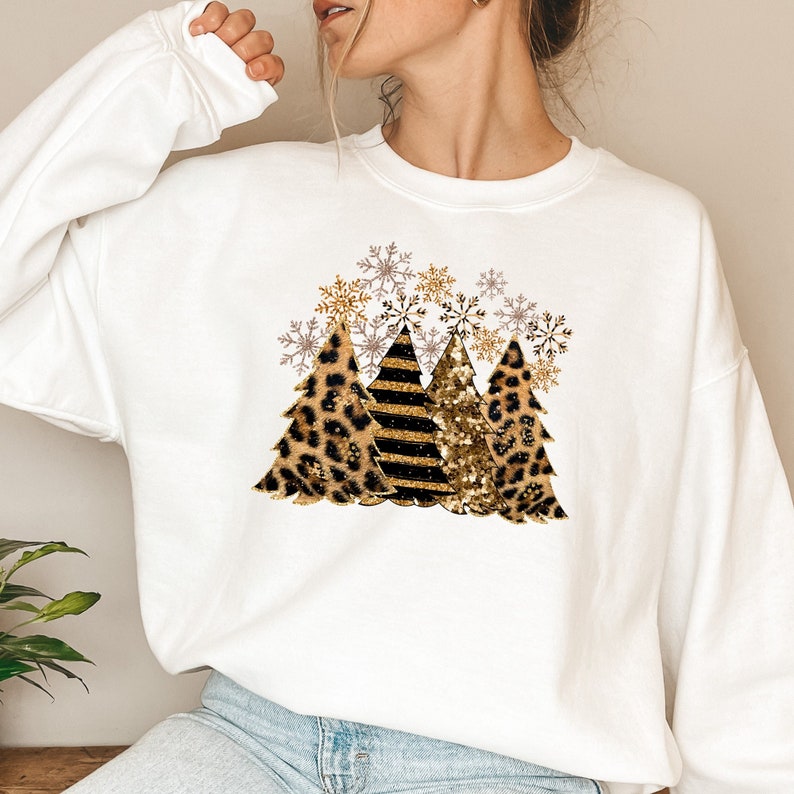 May include: White sweatshirt with a graphic of three Christmas trees. The trees are decorated with leopard print, black and gold stripes, and gold glitter. There are snowflakes around the trees.
