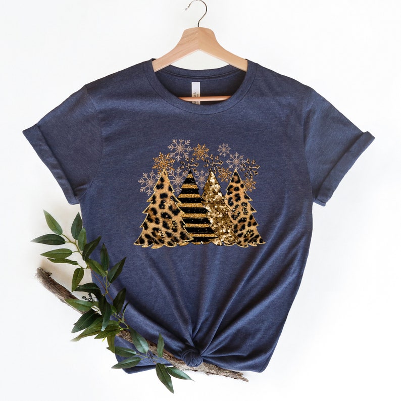 May include: A navy blue t-shirt with a design of three Christmas trees. The trees are decorated with gold glitter, black stripes, and leopard print. Snowflakes are scattered around the trees.